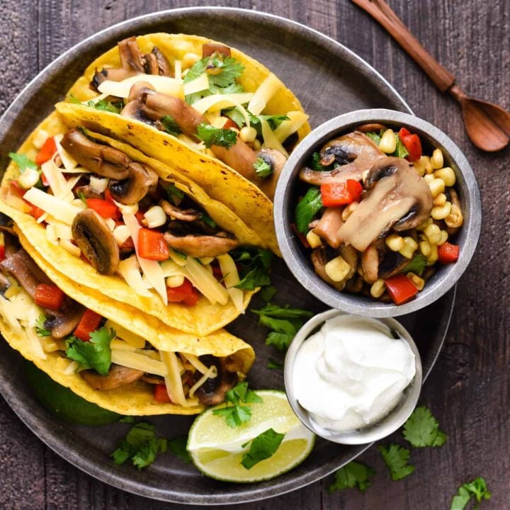 A round tray filled with three mushroom tacos made in corn tortillas, a lime wedge, and small bowls of mushroom filling and sour cream.