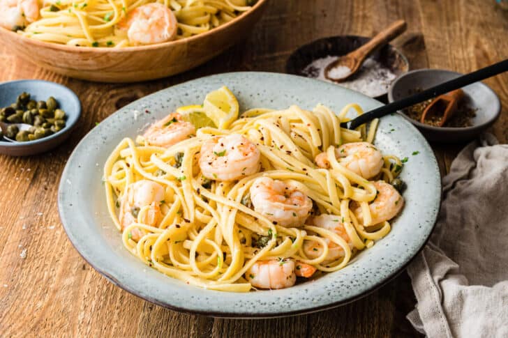 A gray bowl filled with lemon caper pasta and shrimp, with spices and capers and a larger serving bowl of pasta garnishing the scene.