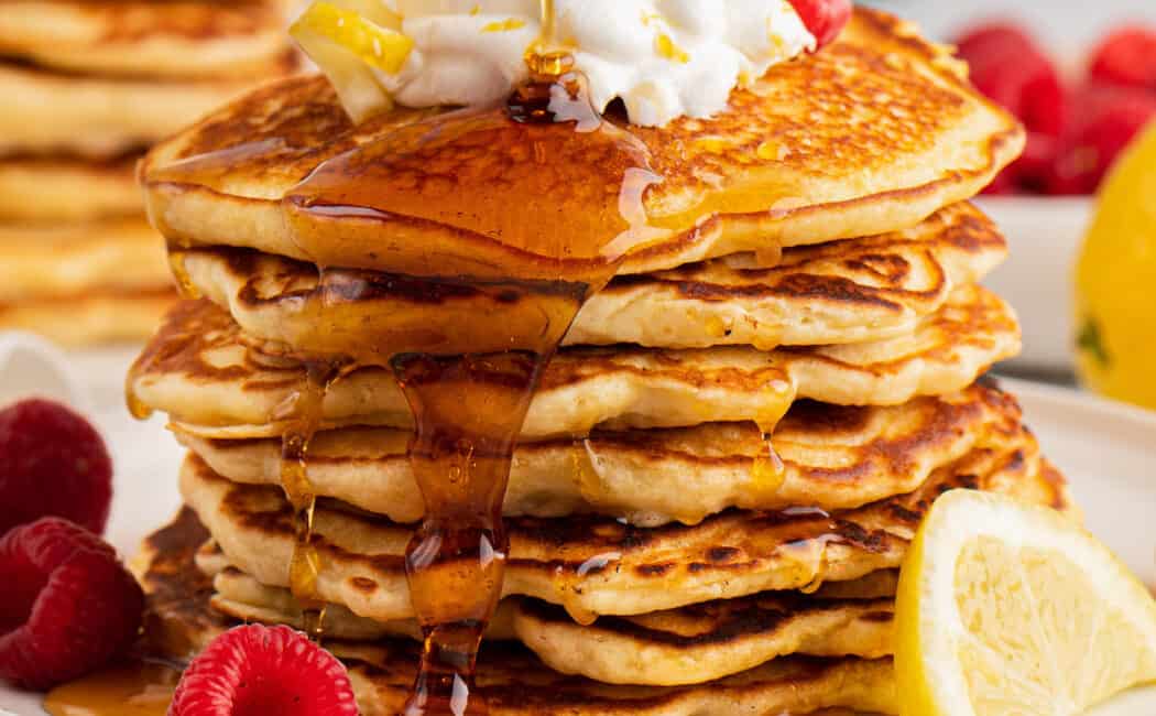 A tall stack of lemon pancakes on a gray plate, garnished with berries and whipped cream, being drizzled with maple syrup.