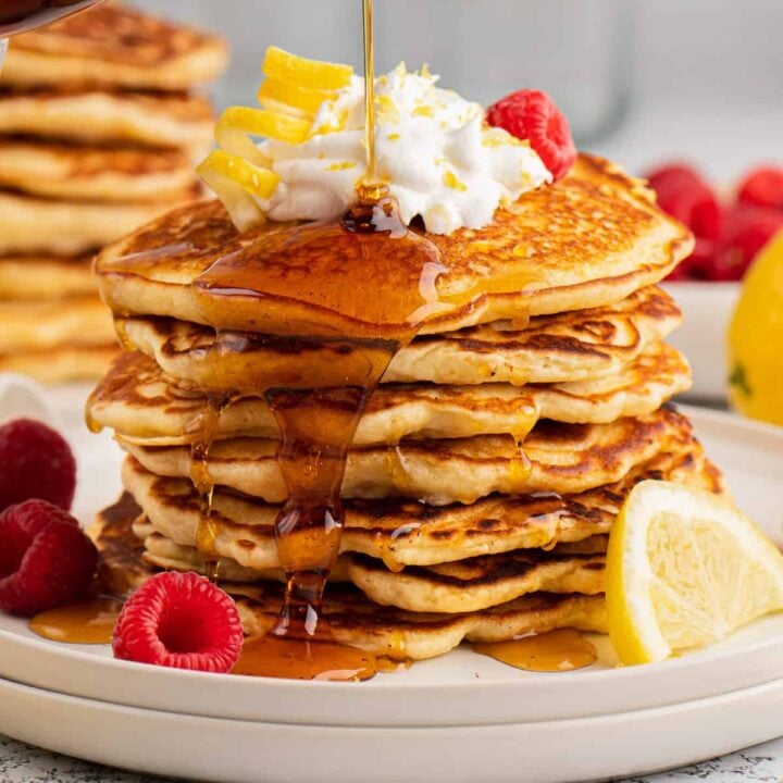 A tall stack of lemon pancakes on a gray plate, garnished with berries and whipped cream, being drizzled with maple syrup.
