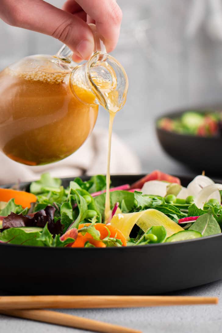 Ginger miso dressing being poured out of a small glass pitcher over a fresh vegetable salad.