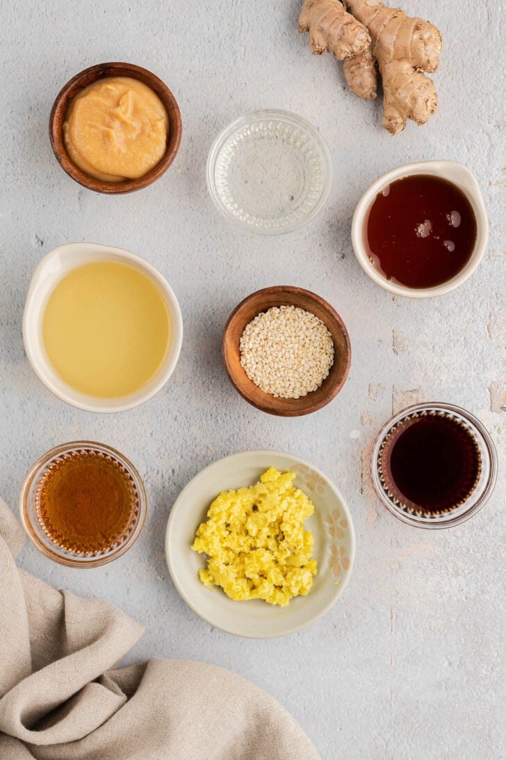 Ingredients on a white surface, including grated ginger, oil, vinegar, sesame seeds, miso and soy sauce.