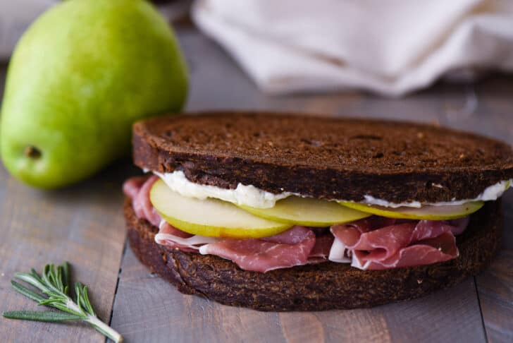 A prosciutto sandwich made with pears and pumpernickel bread on a rustic wooden tabletop.
