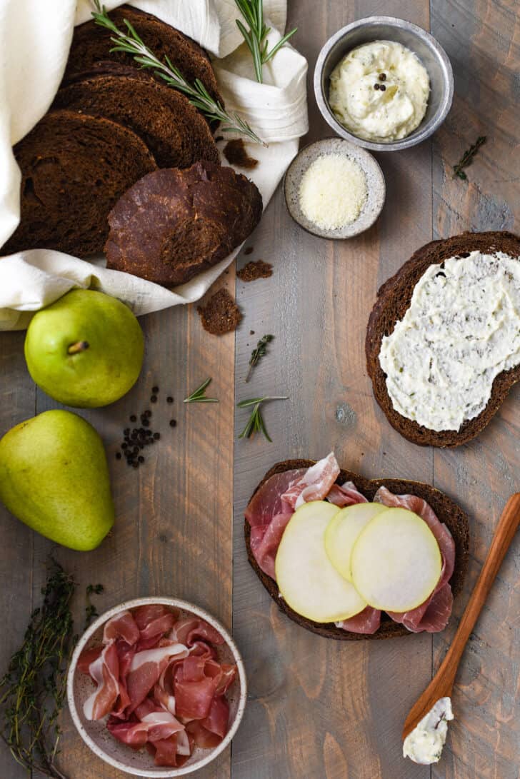 The makings for a prosciutto sandwich recipe on a rustic wooden background, including bread, pears, mayonnaise, cheese and herbs.