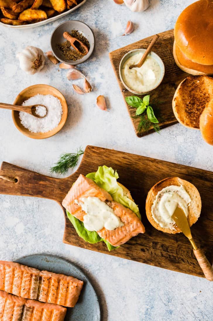 Fixings for sandwiches, including brioche buns, grilled salmon, herb mayo, lettuce and herbs on small cutting boards on a tabletop.
