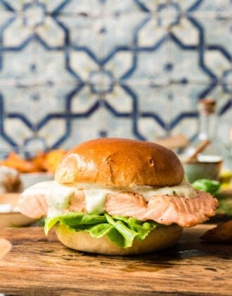 A grilled salmon sandwich with lettuce and mayo on a brioche hamburger bun, on a small cutting board in front of a colorful tile backsplash.