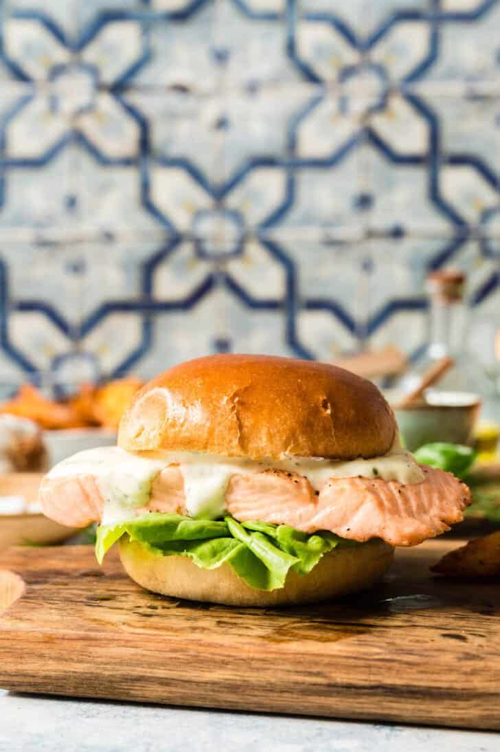 A grilled salmon sandwich with lettuce and mayo on a brioche hamburger bun, on a small cutting board in front of a colorful tile backsplash.