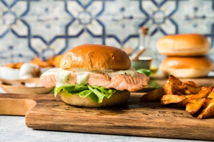 Grilled fish with lettuce and mayo on a brioche hamburger bun, on a small cutting board with a pile of roasted potatoes, in front of a colorful tile backsplash.