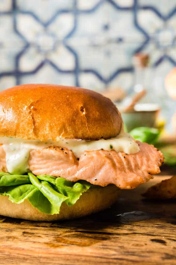 A grilled salmon sandwich with lettuce and mayo on a brioche hamburger bun, on a small cutting board in front of a colorful tile backsplash.