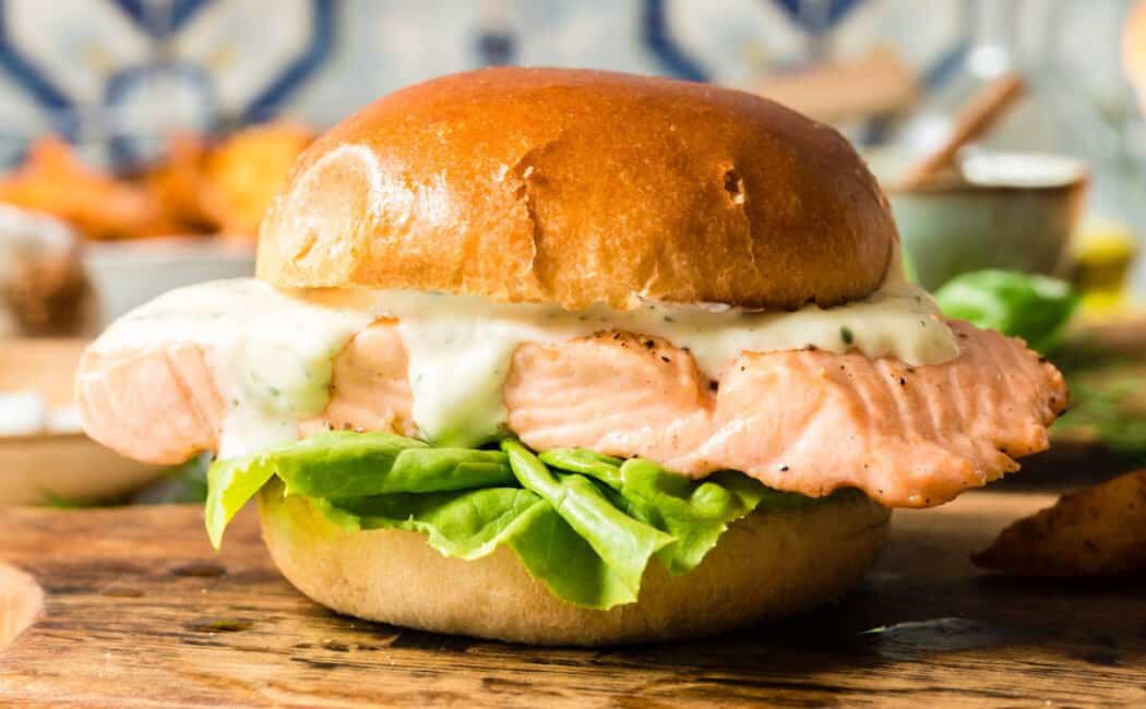 A grilled salmon sandwich with lettuce and mayo on a brioche hamburger bun, on a small cutting board in front of a colorful tile backsplash.