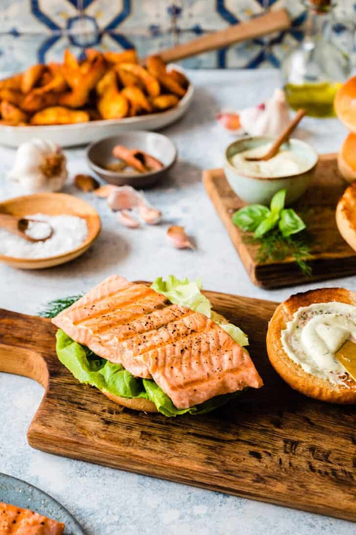 A piece of grilled fish on top of lettuce and a bun on a small wooden cutting board.