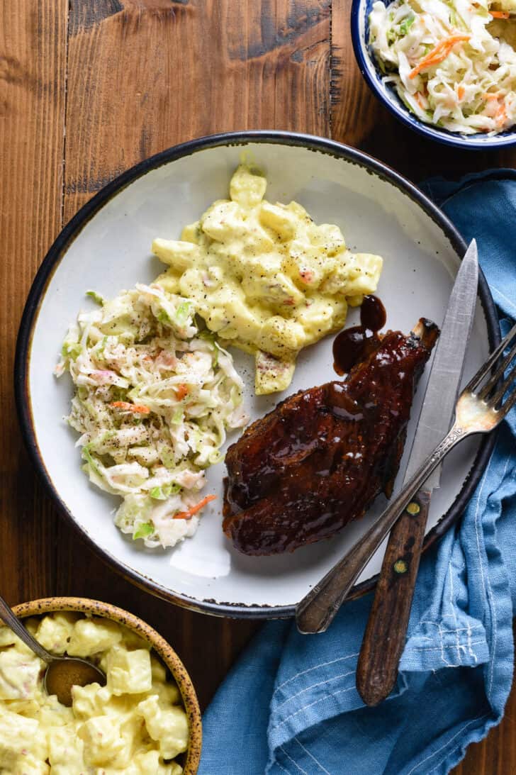 A rustic plate filled with potato salad, coleslaw and a country style pork rib with barbecue sauce.