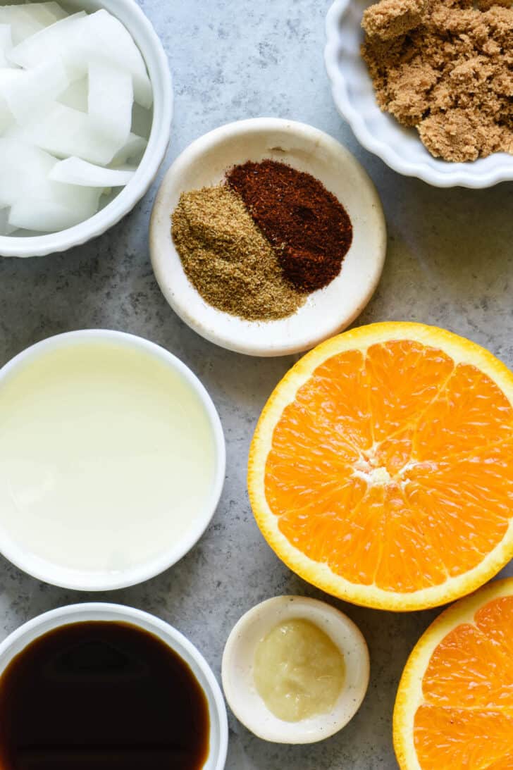 Ingredients for a flat iron steak marinade on a light colored surface, including spices, brown sugar, chopped onions, halved oranges, oil, soy sauce and garlic paste.