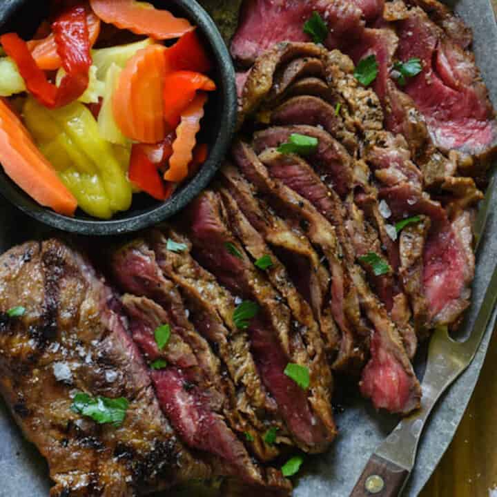 Grilled flat iron steak on a serving platter with a meat fork, pickled vegetables and sea salt.