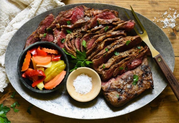 An oval steel platter topped with a sliced grilled flat iron steak and garnishes.