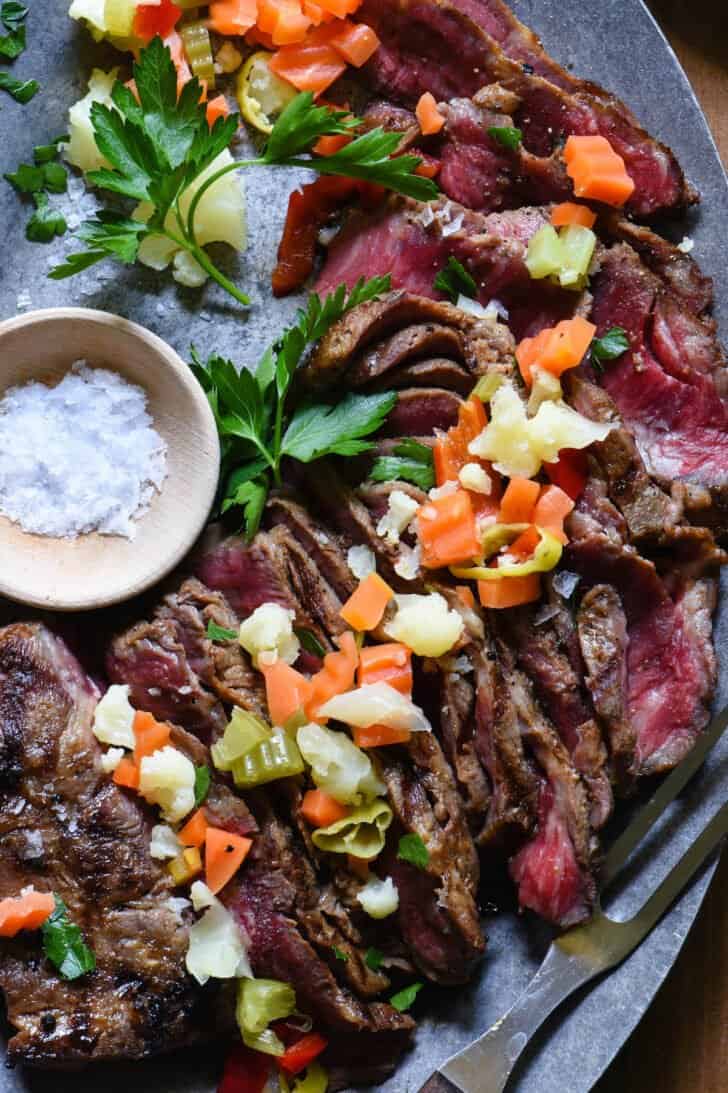 An oval steel platter topped with a sliced flat iron steak on grill and garnishes.