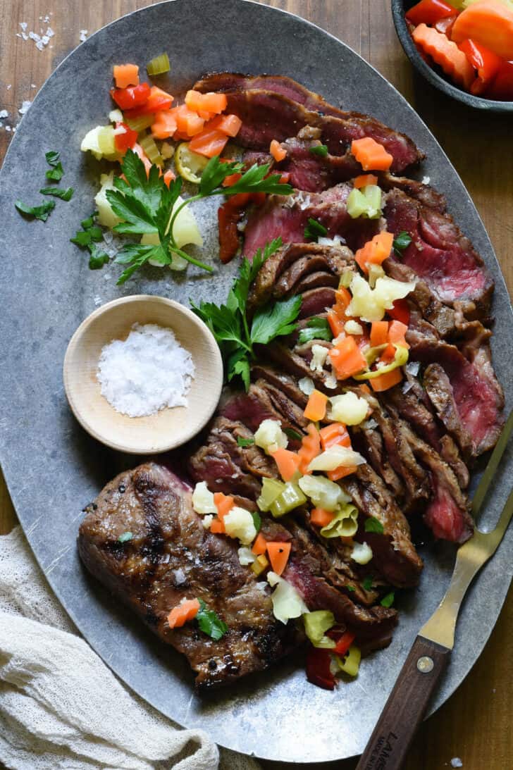 Grilled flat iron steak on a serving platter with a meat fork, pickled vegetables and sea salt.
