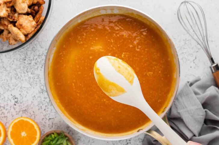A white spoon showing the thickness of an orange sauce in a skillet.