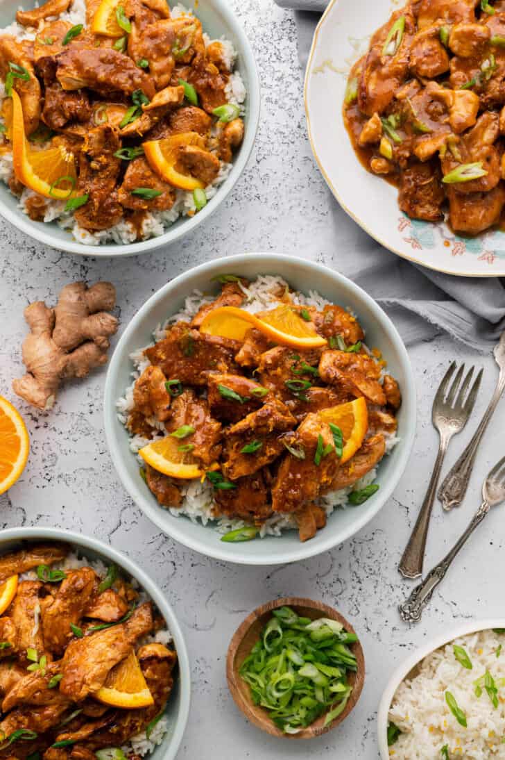 Several bowls and plates of homemade orange chicken served over rice on a light surface.