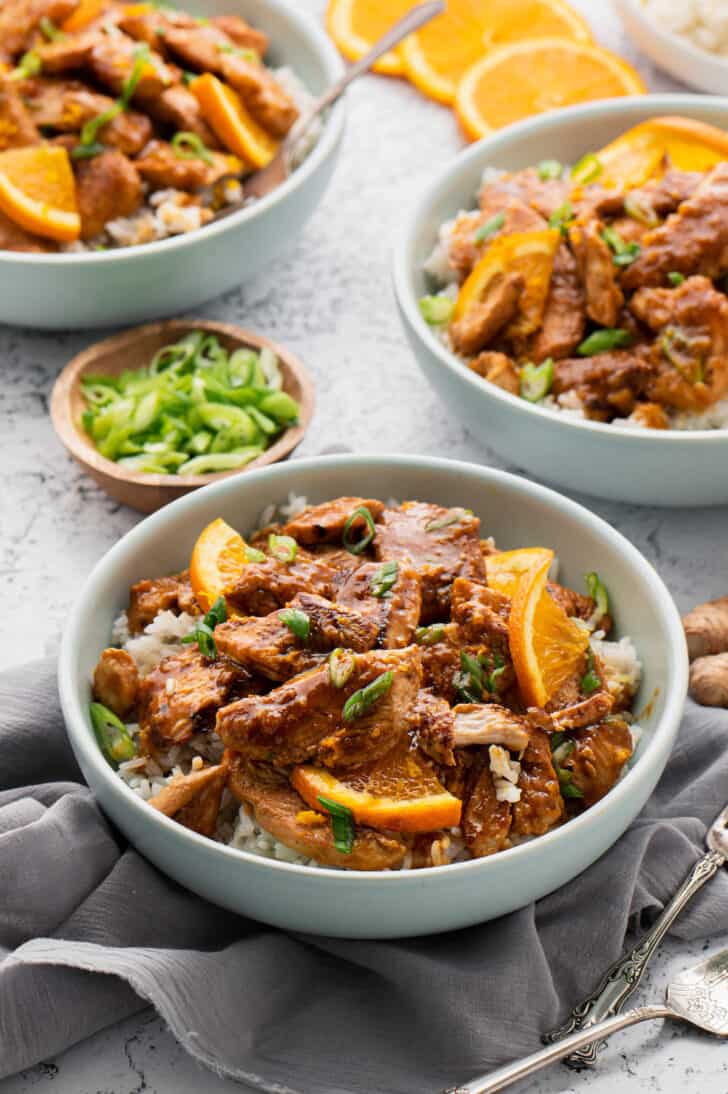 White bowls filled with rice and homemade orange chicken, garnished with orange slices and chopped green onions.