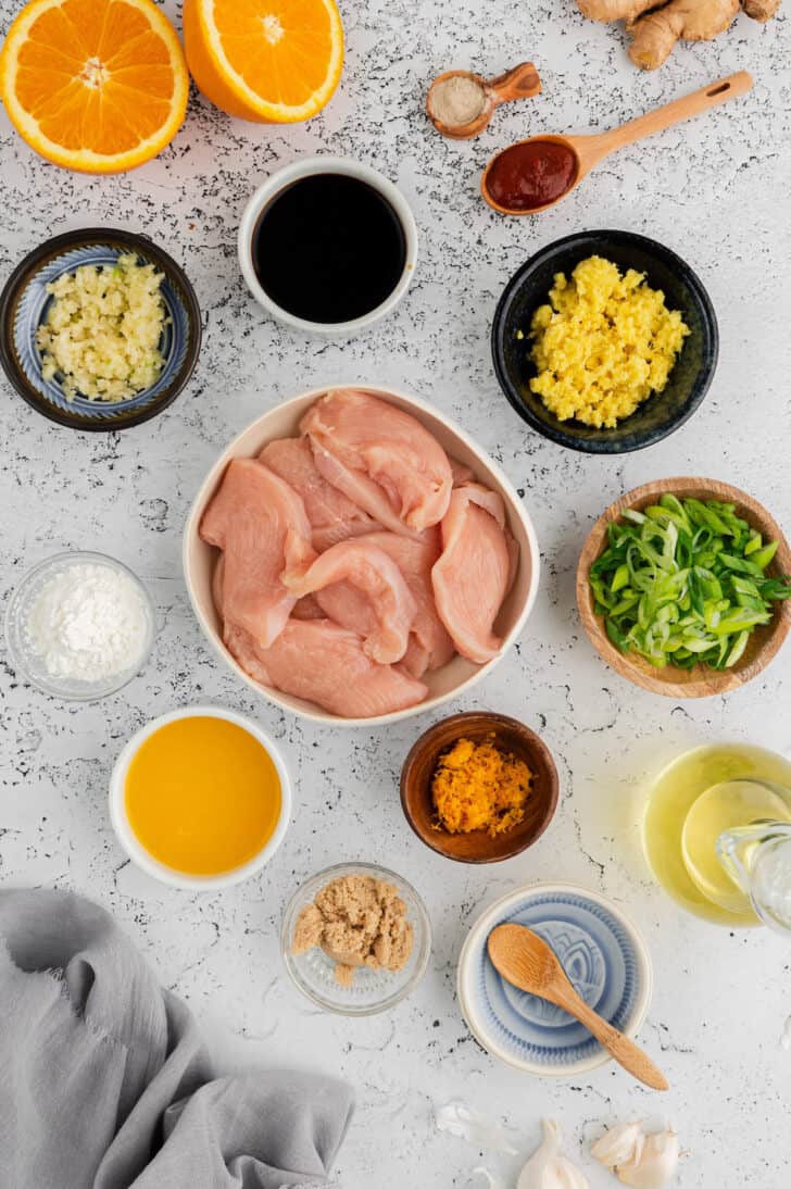 Ingredients for spicy orange chicken laid out on a light surface, including chicken, garlic, ginger, Asian condiments and oranges.