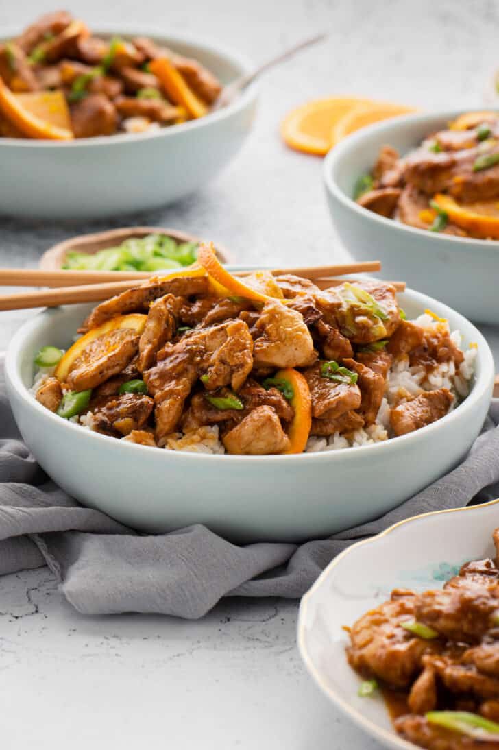 White bowls filled with rice and chicken with orange chicken sauce with a set of chopsticks taking out a bite.