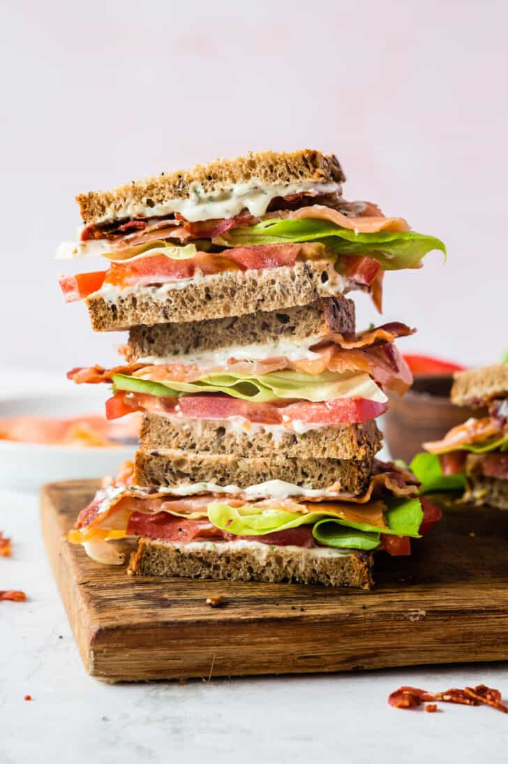 A stack of smoked salmon BLT sandwich halves on a wooden cutting board in front of a light pink backdrop.