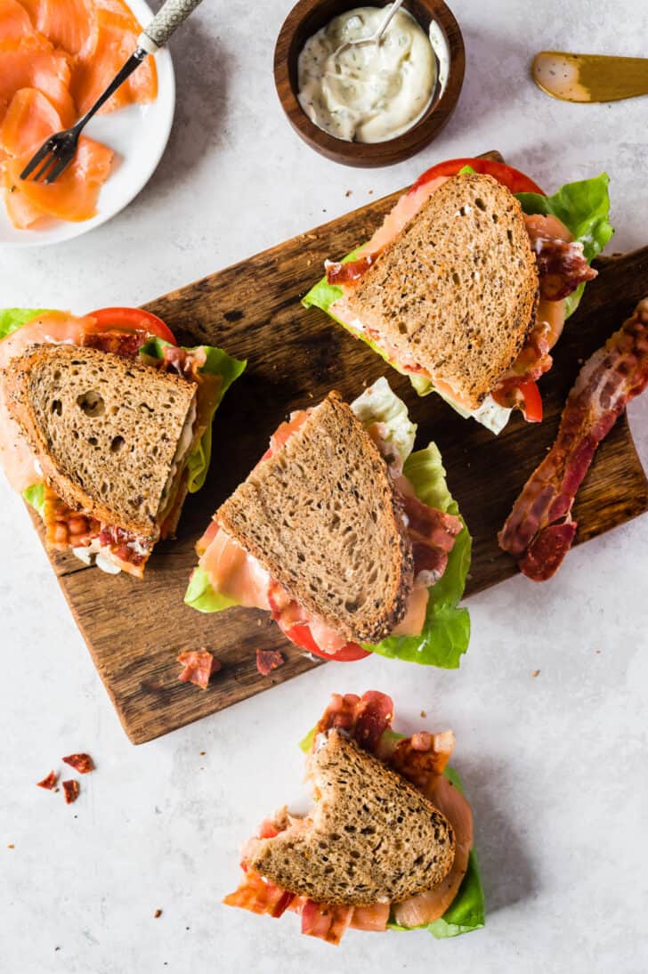 Salmon BLT sandwich halves on a wooden cutting board on a pale pink tabletop.