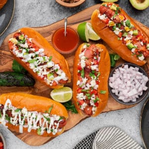 A rustic cutting board topped with Sonoran hot dogs, limes, onions and hot sauce.