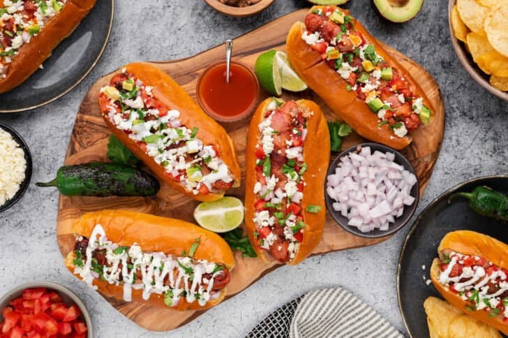 A rustic cutting board topped with Mexican hot dogs, limes, onions and hot sauce.