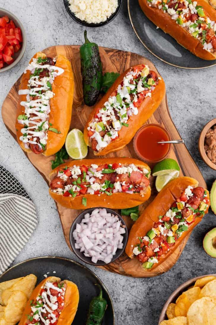 A rustic cutting board topped with Sonoran hot dogs, limes, onions and hot sauce.