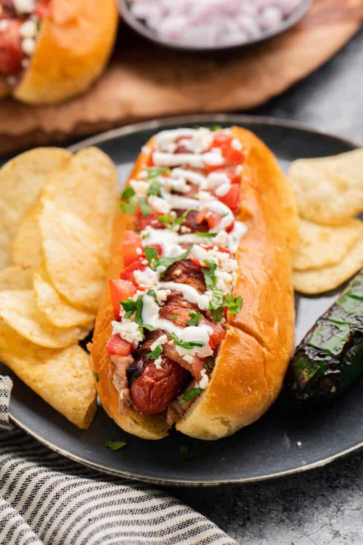 Bacon wrapped hot dog topped with sour cream, tomatoes, cilantro and cheese, on a black plate with potato chips.