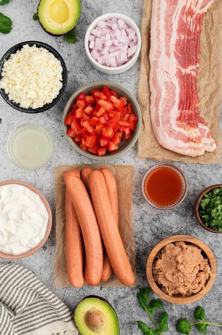 The ingredients for Mexican street hot dogs laid out on a light surface, including hot dogs, bacon, tomatoes, herbs, cheese, refried beans, sour cream and avocado.