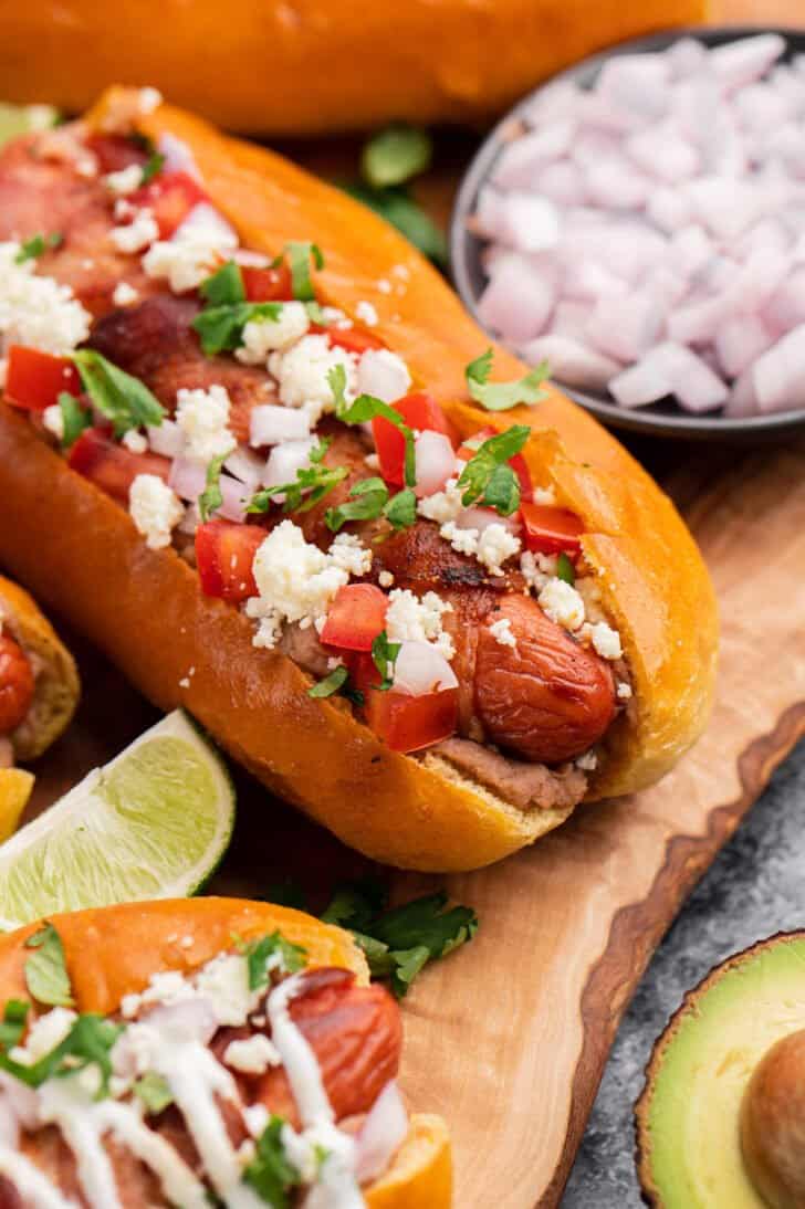 Closeup on a bacon wrapped Sonoran hot dog topped with cheese, tomatoes, herbs and onions.