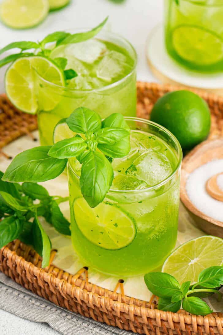 Bright green cocktails with basil on a wicker gray with limes and fresh basil garnishing the tray.