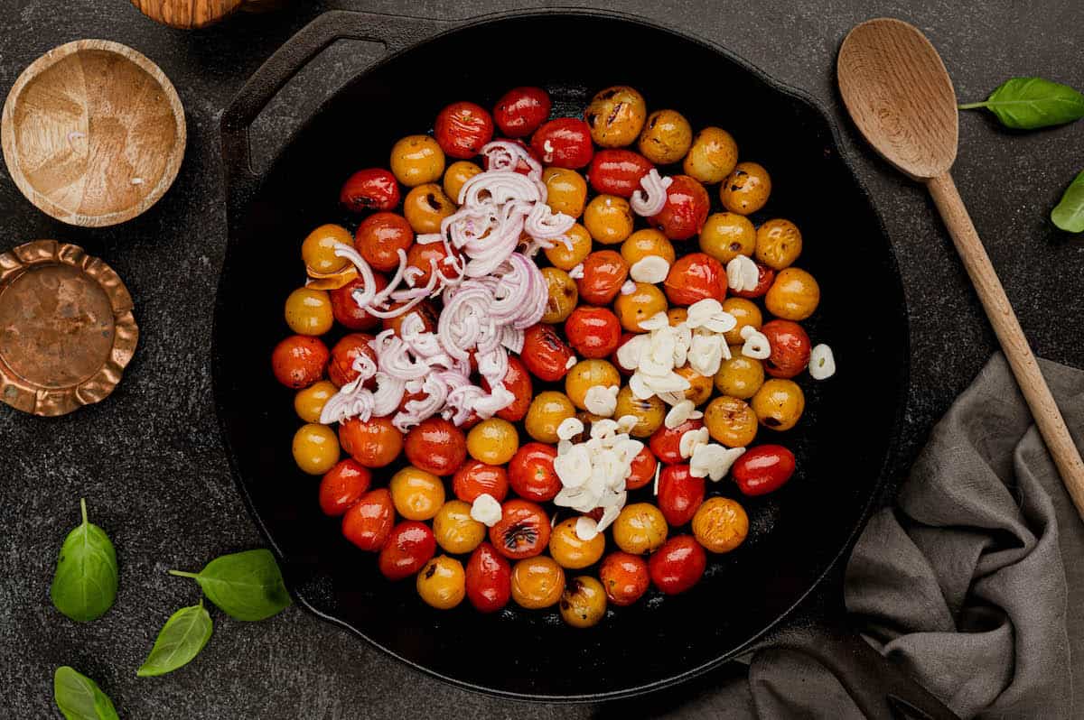 Blister tomatoes in a cast iron skillet topped with sliced shallot and garlic.