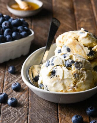 Small oval dish filled with scoops of blueberry cheesecake ice cream, with a bowl of blueberries and a dish of honey in the background.