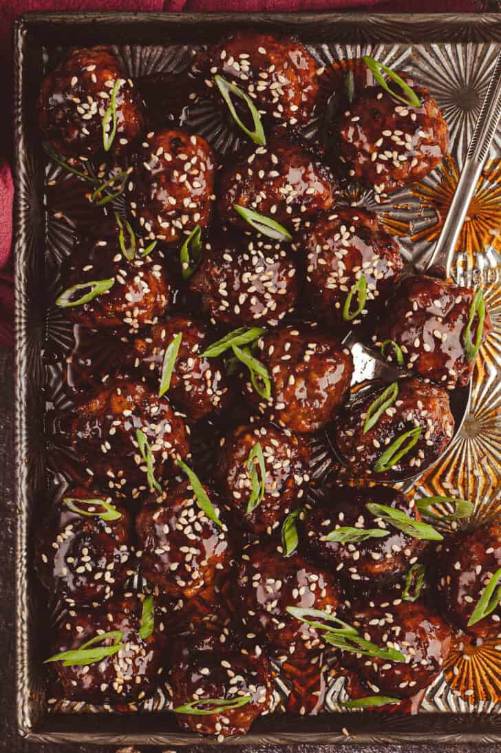 Korean meatballs glazed with a deep red sauce on a textured baking pan, garnished with sliced green onions and white sesame seeds.