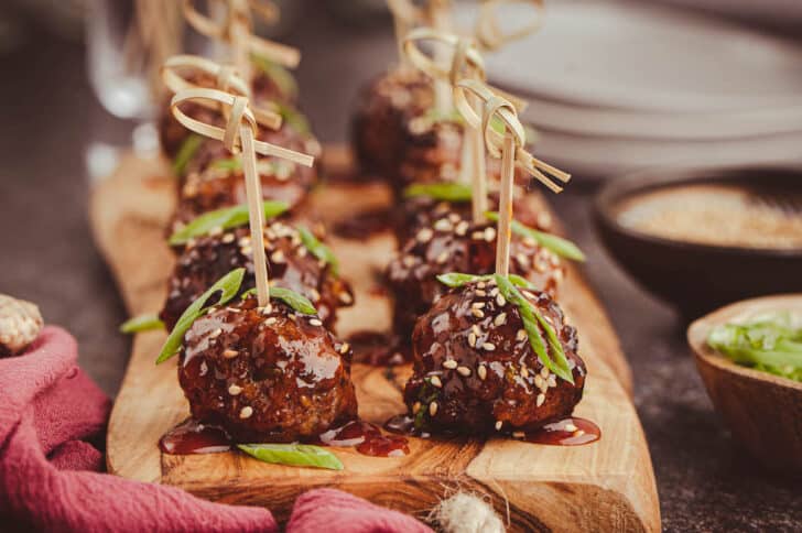 Saucy meatballs on a small wooden cutting board with decorative wooden toothpicks skewered into each one, garnished with green onions and sesame seeds.