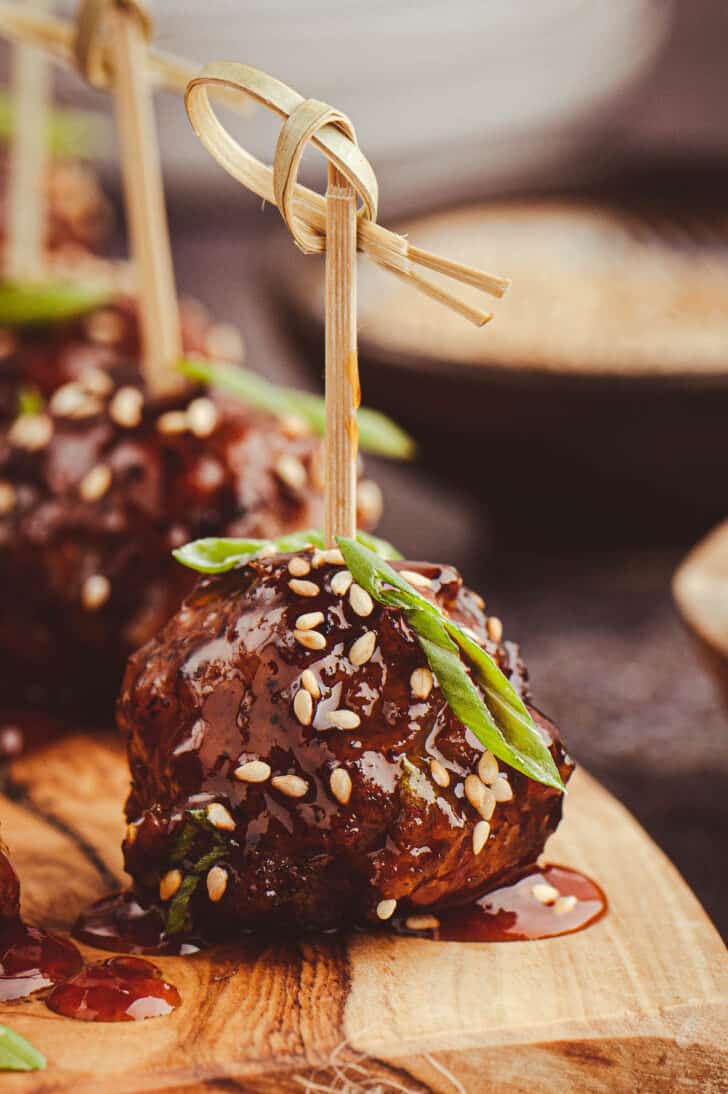 A closeup on a Korean meatball skewered with a decorative toothpick and garnished with sesame seeds and green onions, on a small wooden cutting board.