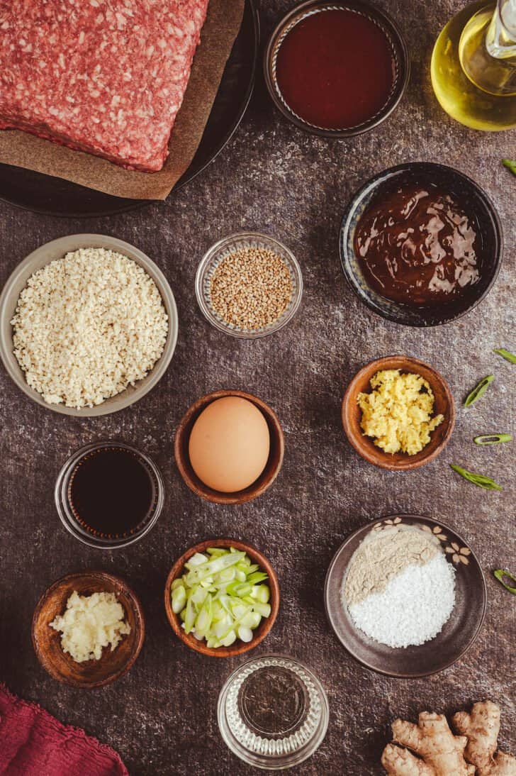 The ingredients for Korean BBQ meatballs on a dark textured surface, including ground beef, egg, and bowls of sauces, breadcrumbs, spices, onions and sesame seeds.