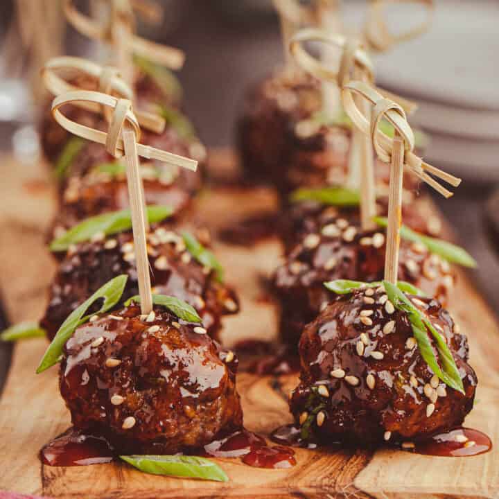 Saucy Korean meatballs on a small wooden cutting board with decorative wooden toothpicks skewered into each one, garnished with green onions and sesame seeds.