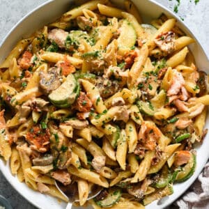 A white skillet with a metal handle on a light gray surface, filled with penne pasta, zucchini, mushrooms and salmon chunks tossed with creamy sauce and sprinkled with green herbs and Parmesan cheese, with a pink linen on the table underneath the skillet handle, and a spoon digging into the skillet.