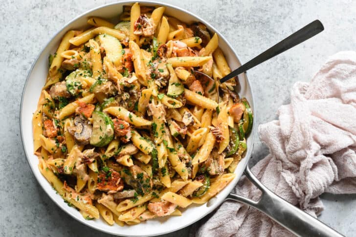 A white skillet with a metal handle on a light gray surface, filled with penne pasta, zucchini, mushrooms and salmon chunks tossed with creamy sauce and sprinkled with green herbs and Parmesan cheese, with a pink linen on the table nearby, and a spoon digging into the skillet.