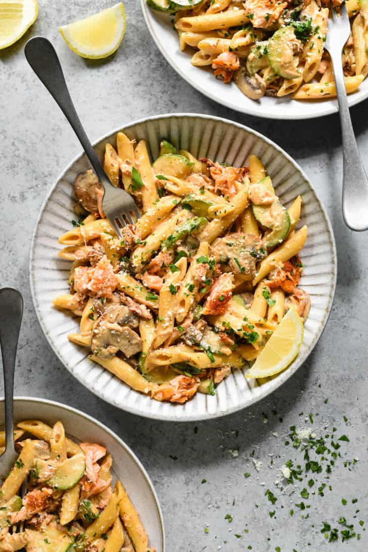 Textured gray ceramic shallow bowl on a light gray surface, filled with Cajun pasta with salmon, topped with parsley and cheese, garnished with a lemon wedge, with a fork digging into it.