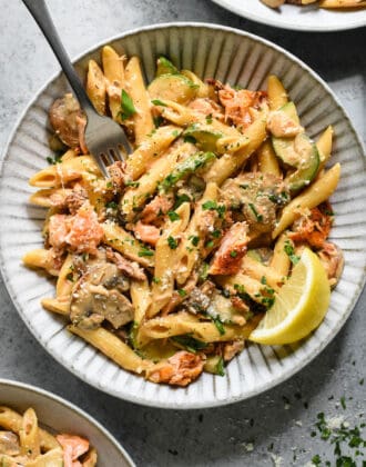 Textured gray ceramic shallow bowl filled with Cajun salmon pasta topped with parsley and cheese, garnished with a lemon wedge, with a fork digging into it.