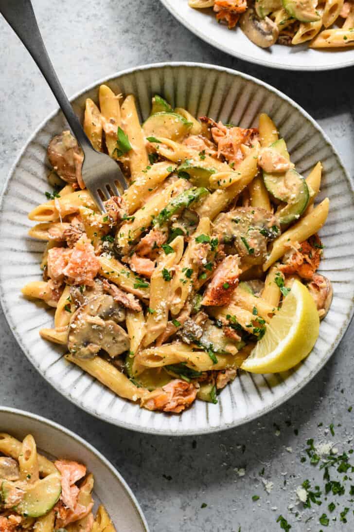 Textured gray ceramic shallow bowl filled with Cajun salmon pasta topped with parsley and cheese, garnished with a lemon wedge, with a fork digging into it.