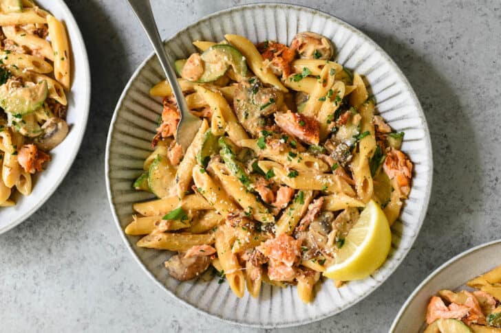 Textured gray ceramic shallow bowl on a light gray surface, filled with penne and pink fish chunks, topped with parsley and cheese, garnished with a lemon wedge, with a fork digging into it.