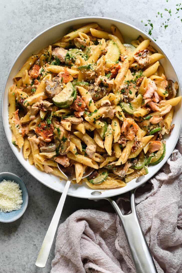 A white skillet with a metal handle on a light gray surface, filled with penne pasta, zucchini, mushrooms and salmon chunks tossed with creamy sauce and sprinkled with green herbs and Parmesan cheese, with a pink linen on the table underneath the skillet handle, and a spoon digging into the skillet.