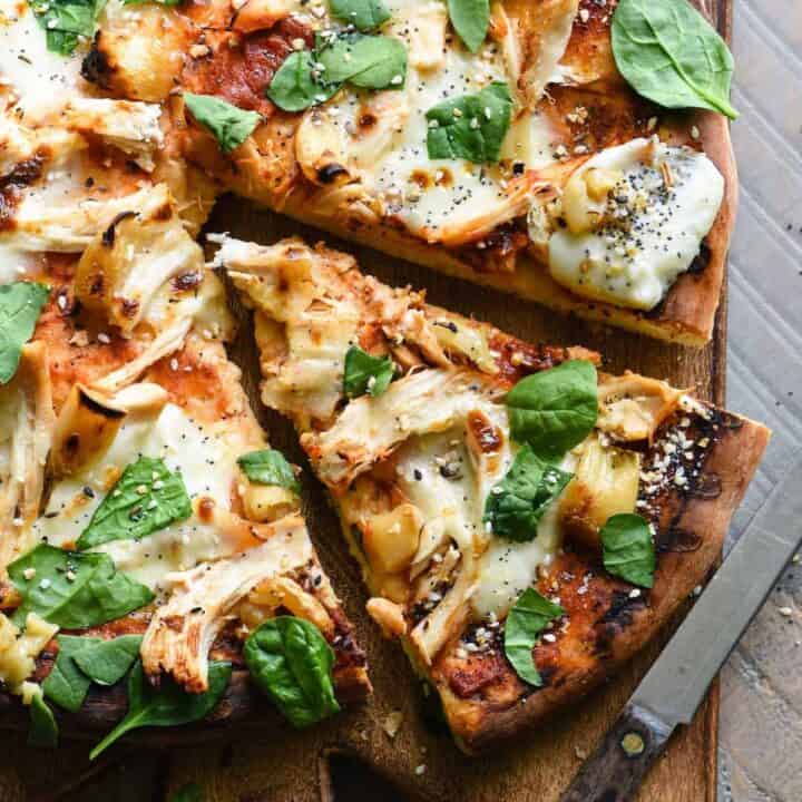 A round chicken pizza with roasted garlic and basil on a wooden cutting board.